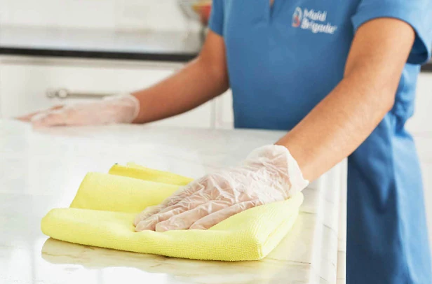 Person in a blue cleaning uniform and disposable gloves wipes a white countertop with a folded yellow cloth.