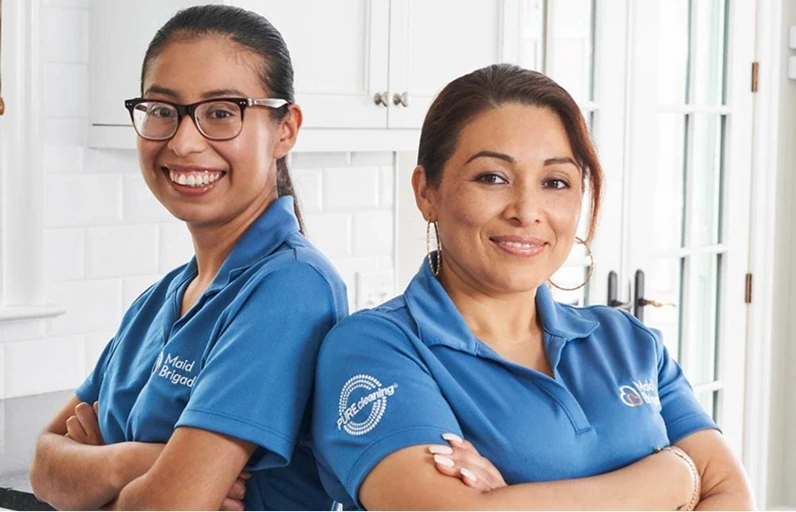 Two cleaning professionals in blue Maid Brigade branded polo shirts stand side by side with arms crossed, smiling in a bright white kitchen; one wears glasses and the other wears hoop earrings.