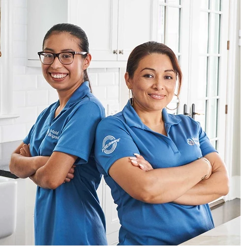 Two cleaning professionals in blue Maid Brigade branded polo shirts stand side by side with arms crossed, smiling in a bright white kitchen; one wears glasses and the other wears hoop earrings.