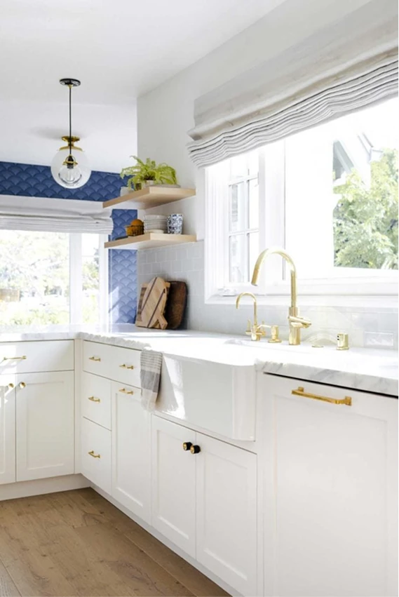 Sunlit white kitchen with shaker cabinets and brass pulls, a white countertop, and a brass faucet beneath a large window with a light Roman shade; blue patterned tile backsplash and floating shelves with plants and decor.
