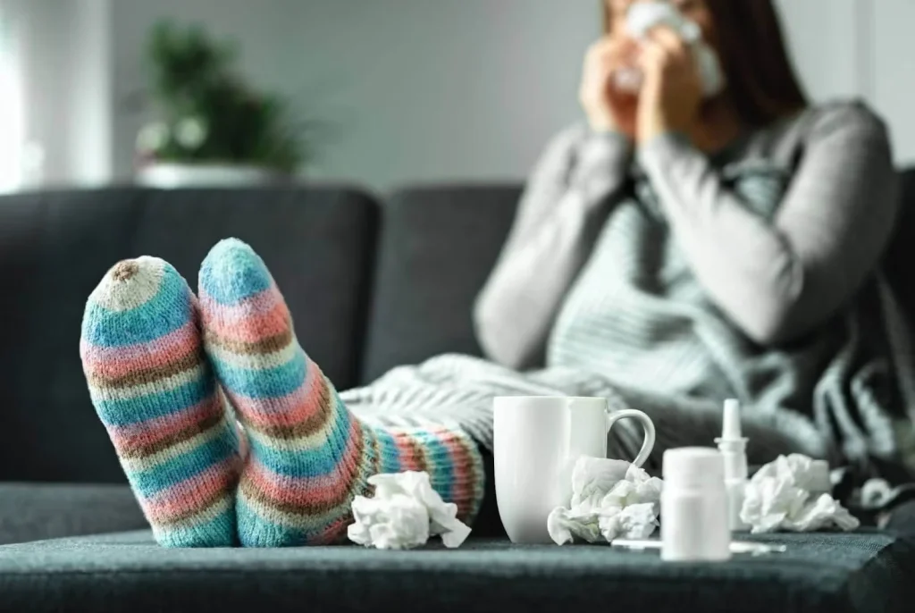 Close-up of colorful striped socks on a couch with tissues, a mug, and medicine in the foreground, while a sick person in the background blows their nose.